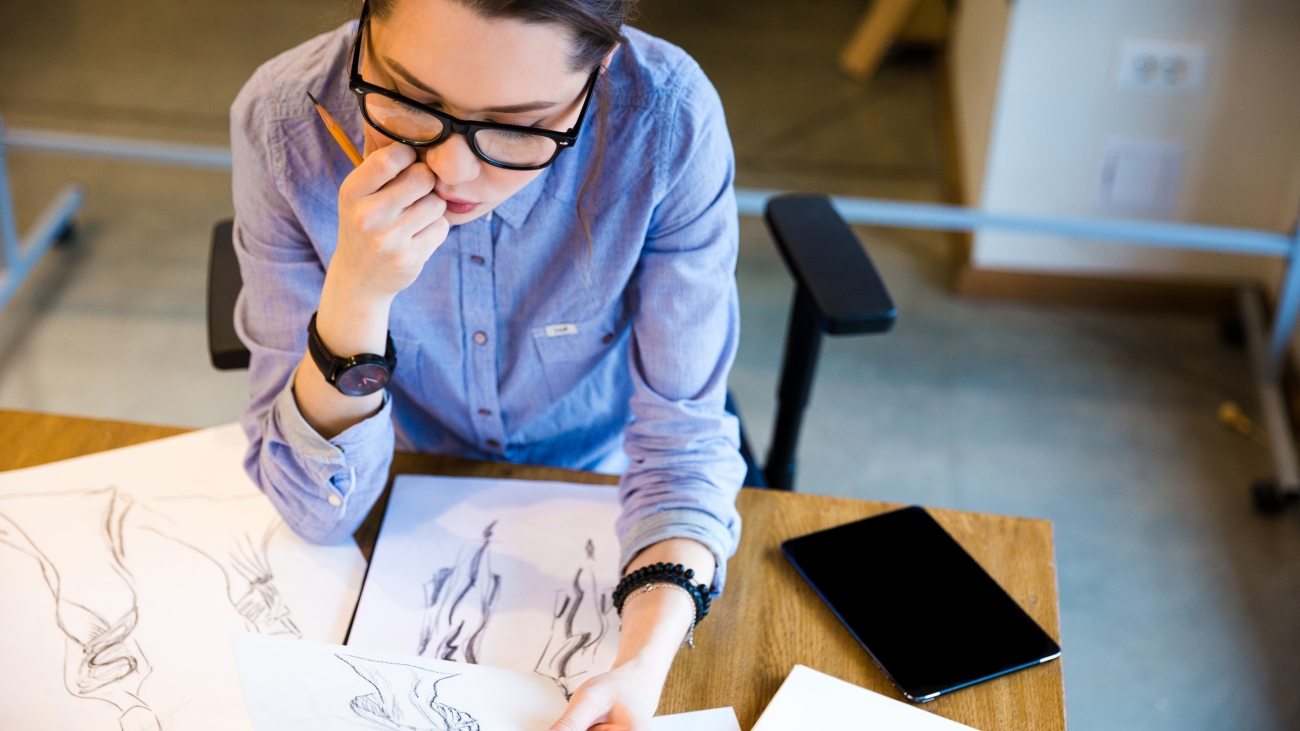 Top view of pensive pretty young woman fashion designer sitting on workplace and looking at sketches