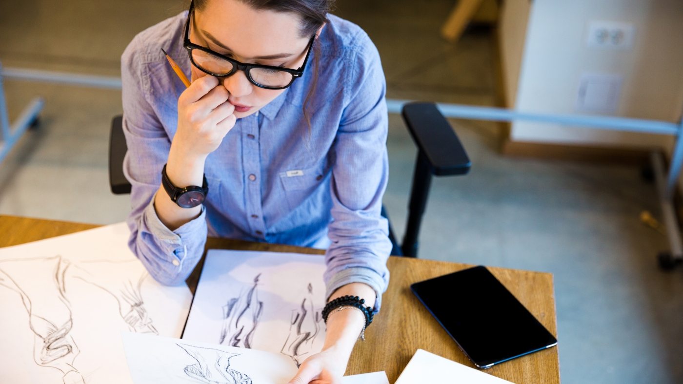 Top view of pensive pretty young woman fashion designer sitting on workplace and looking at sketches