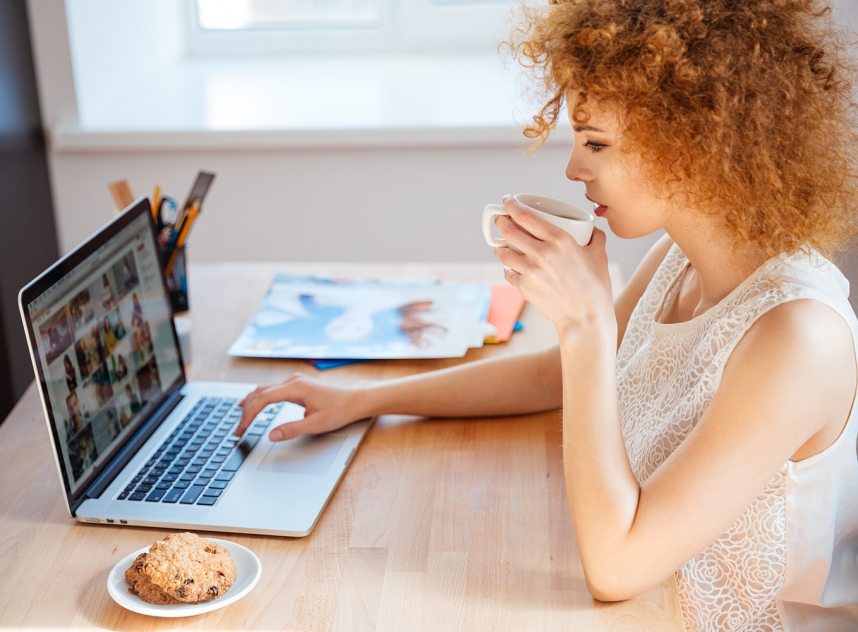 Profile of attractive charming curly young woman photographer drinking coffee and working with laptop on workplace