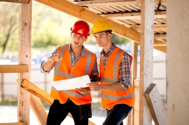 Two men dressed in shirts, orange work vests and helmets explore construction documentation on the building site near the wooden building constructions .