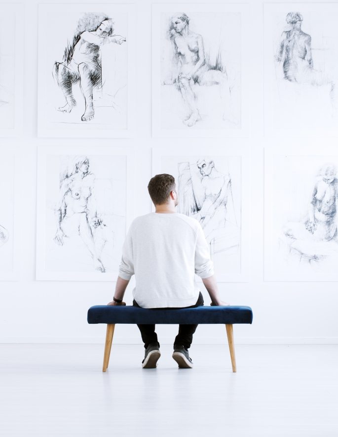 Man relaxing in art gallery while sitting on stool in front of white wall with drawings. Art gallery concept