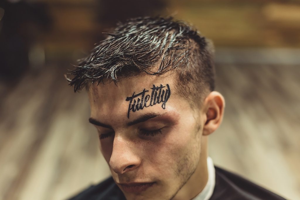 Close-up shot of well-groomed man with hairstyle and tattoo above eyebrow posing with eyes closed.