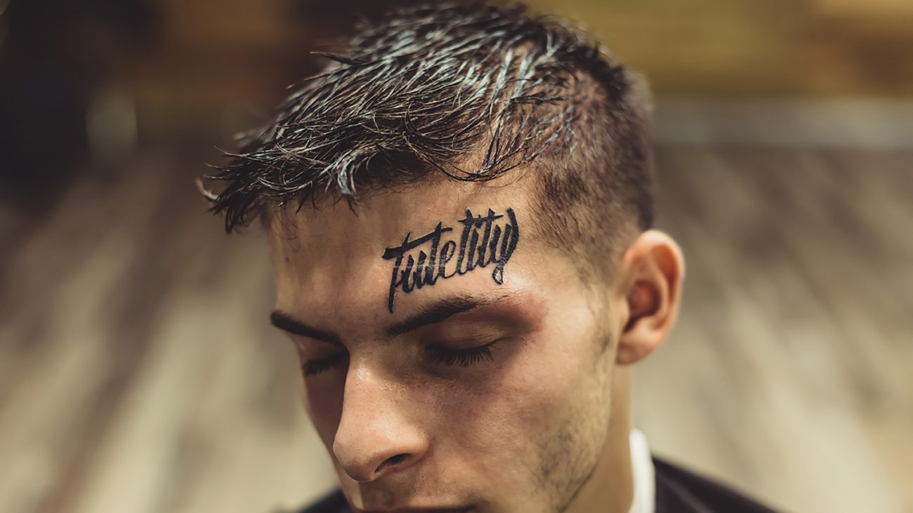 Close-up shot of well-groomed man with hairstyle and tattoo above eyebrow posing with eyes closed.