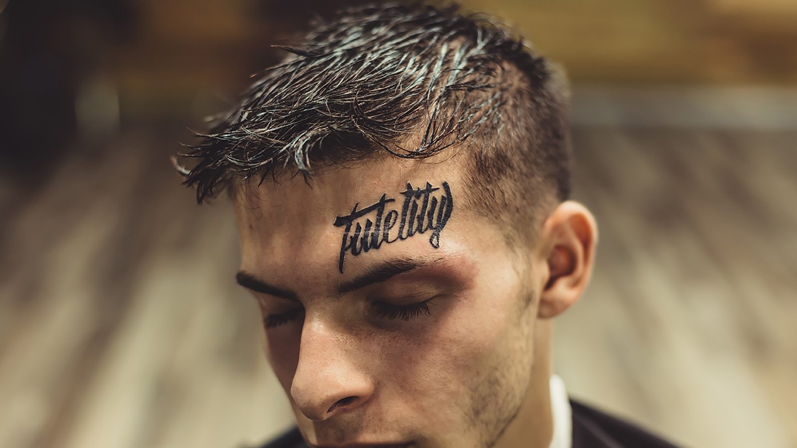 Close-up shot of well-groomed man with hairstyle and tattoo above eyebrow posing with eyes closed.